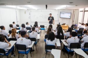 Imagem mostra alunos em uma sala de aula com o professor na frente do quadro de estudos