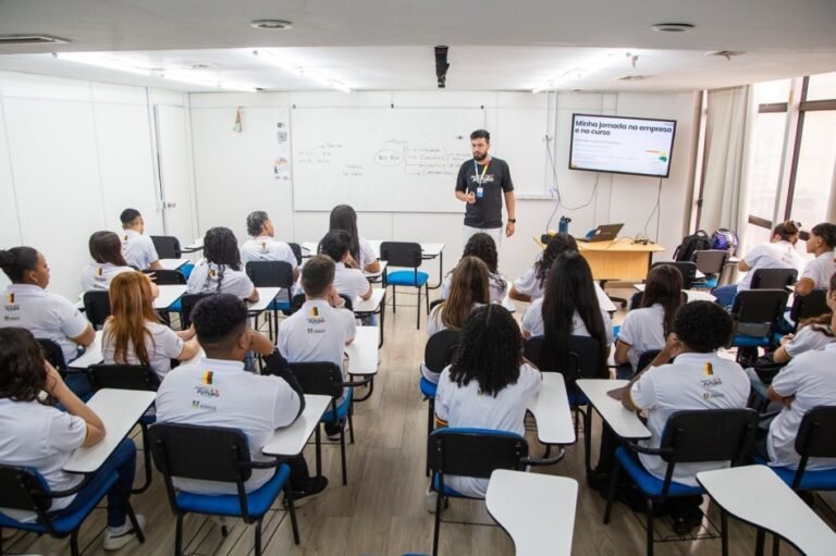 Imagem mostra alunos em uma sala de aula com o professor na frente do quadro de estudos